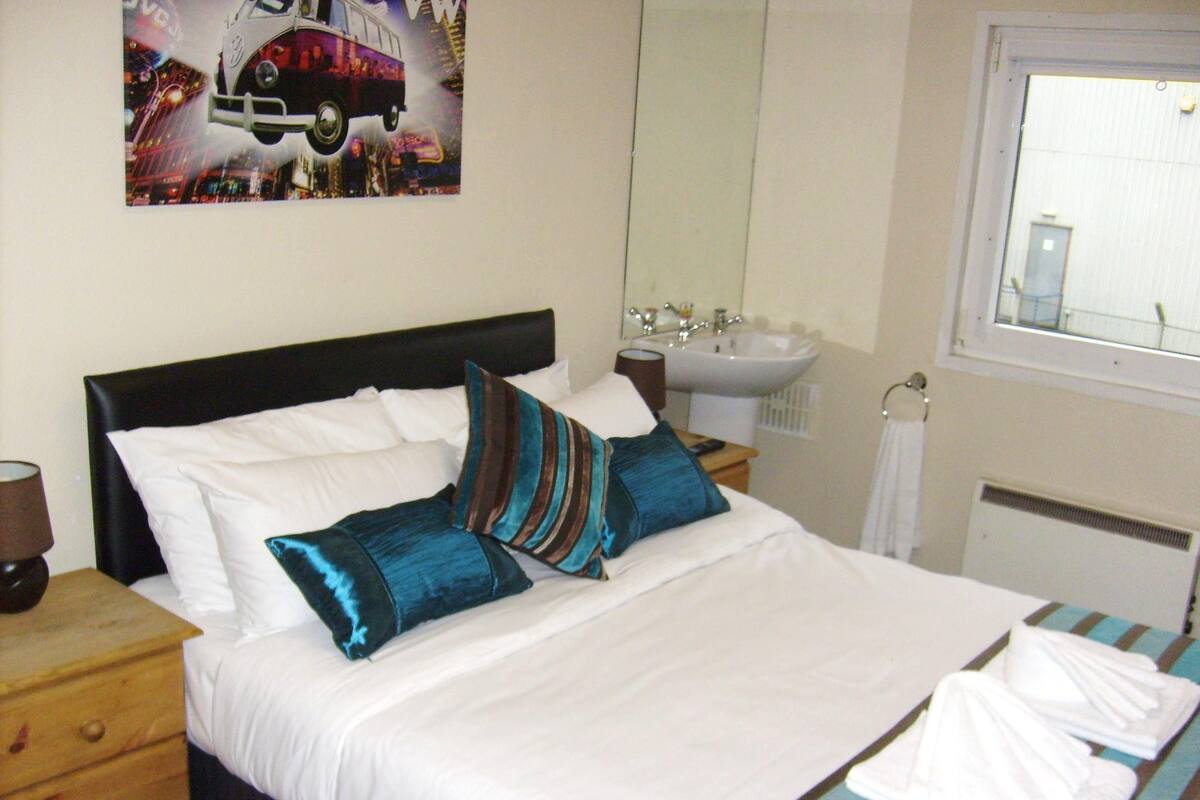 A neatly made bed with white and blue pillows, a wooden nightstand, a bedside lamp, a bathroom with a sink and mirror, and a window with a view of the outside.