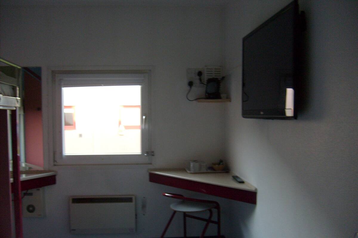 A small, dimly lit room with a single bed, a desk with a chair, a radiator, and a window with a view of buildings outside.