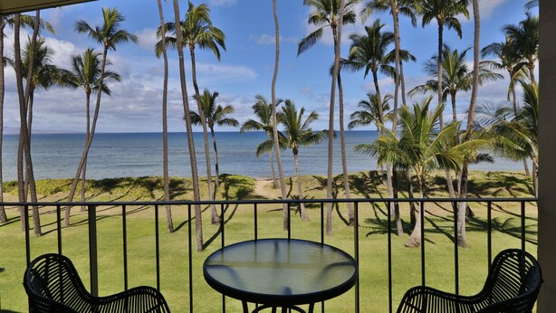 A balcony with a glass table and two chairs, overlooking a grassy area with palm trees and a view of the ocean. - Updated unit, modern and clean (Kihei)