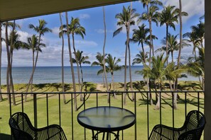 A balcony with a glass table and two chairs, overlooking a grassy area with palm trees and a view of the ocean. - Updated unit, modern and clean (Kihei)