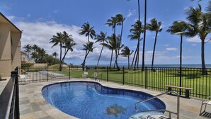 A swimming pool with a curved design, surrounded by a stone deck and a metal fence. - Updated unit, modern and clean (Kihei)