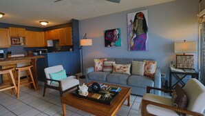 A living room with a coffee table, two chairs, a sofa, and a dining area with wooden cabinets and a microwave. - Updated unit, modern and clean (Kihei)