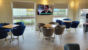 A modern interior with a TV mounted on the wall, blue and beige chairs, wooden tables, and large windows offering a view of the outside.