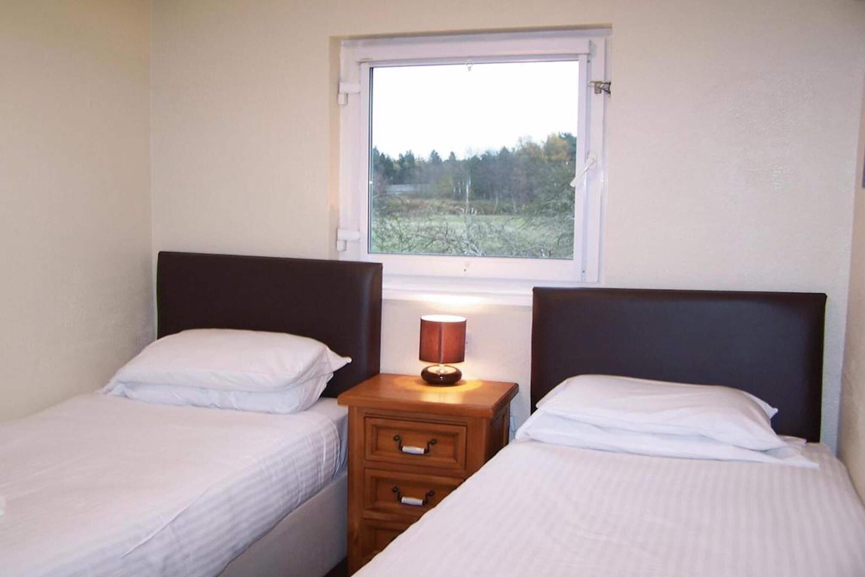 Una habitación con dos camas individuales, una mesita de noche de madera con una lámpara, y una ventana con vistas a árboles.