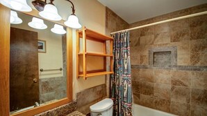 A bathroom with a double sink, a toilet, a bathtub, a mirror with lights, and a shower curtain.