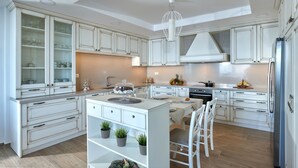 A modern kitchen with white cabinetry, a central island, and a dining area with white chairs. - Villa Omikron (Malevizi)