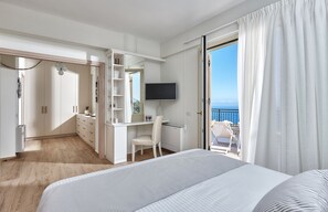 A hotel room with a bed, a desk, a chair, a TV, and a balcony with a view of the sea.