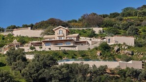A hillside property with multiple buildings, including a central house with a prominent tower, surrounded by lush greenery and trees. - Villa Omikron (Malevizi)