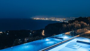 A nighttime view of a coastal city with a pool area featuring glass railings and steps leading into the water. - Villa Omikron (Malevizi)