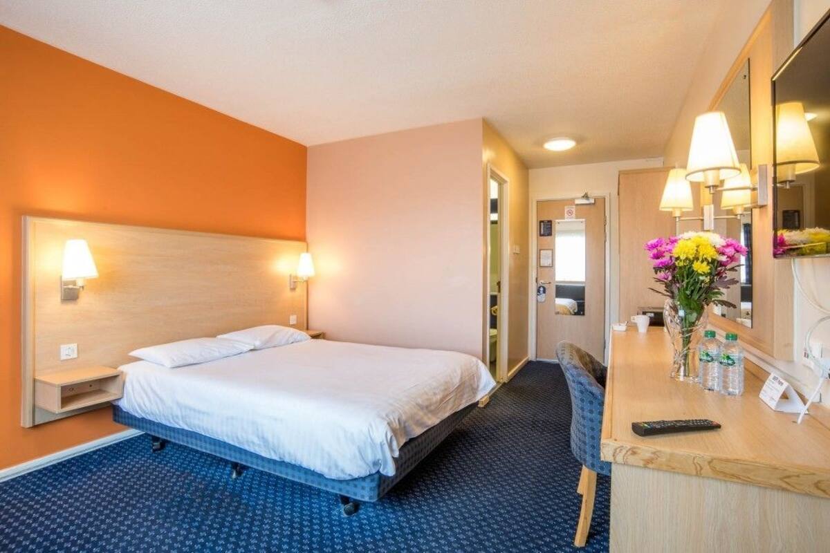 A hotel room with a bed, bedside table, lamp, mirror, and a small desk with a chair.