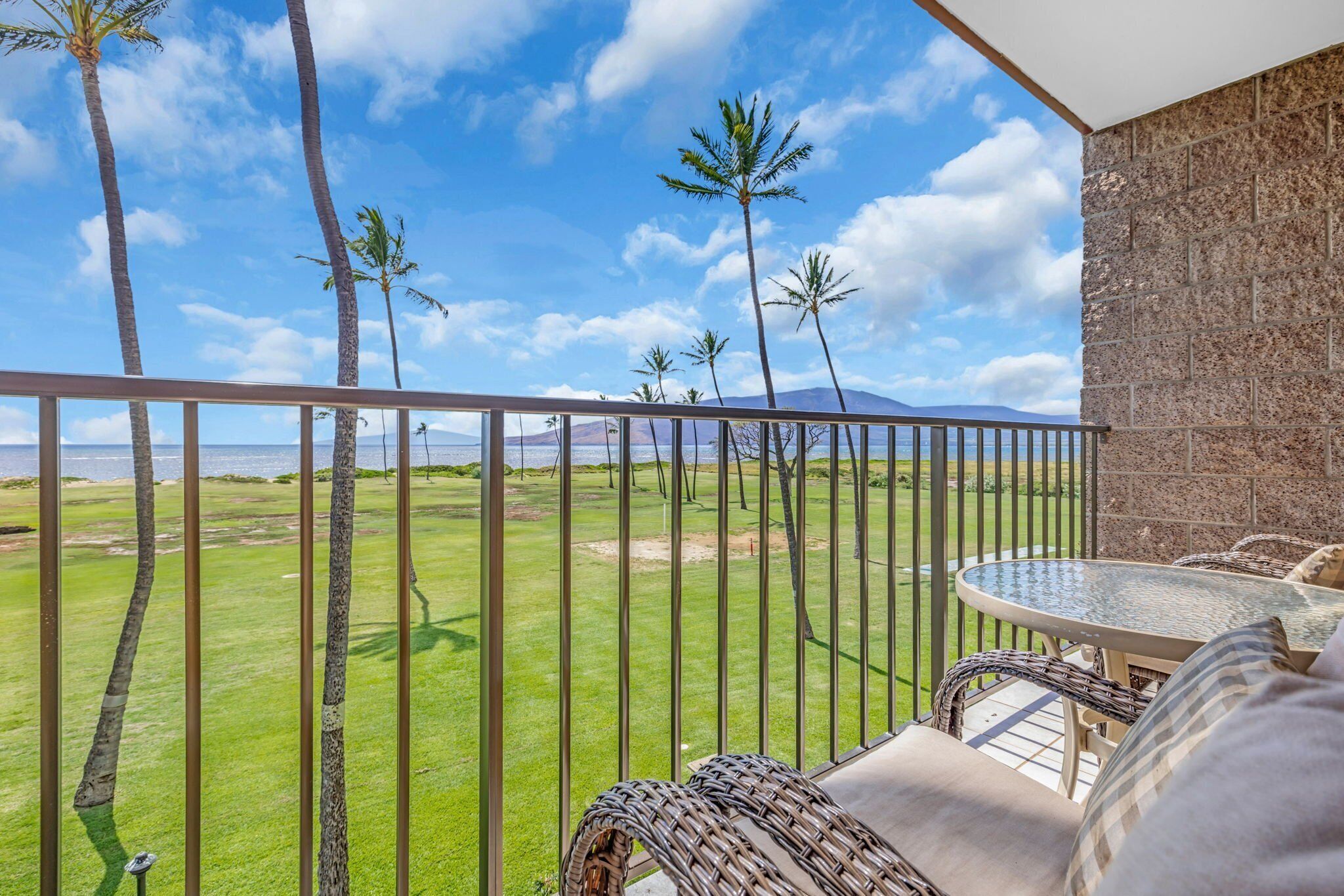Un balcón con vista a un área verde y palmeras, una silla de mimbre y una mesa con cojines.