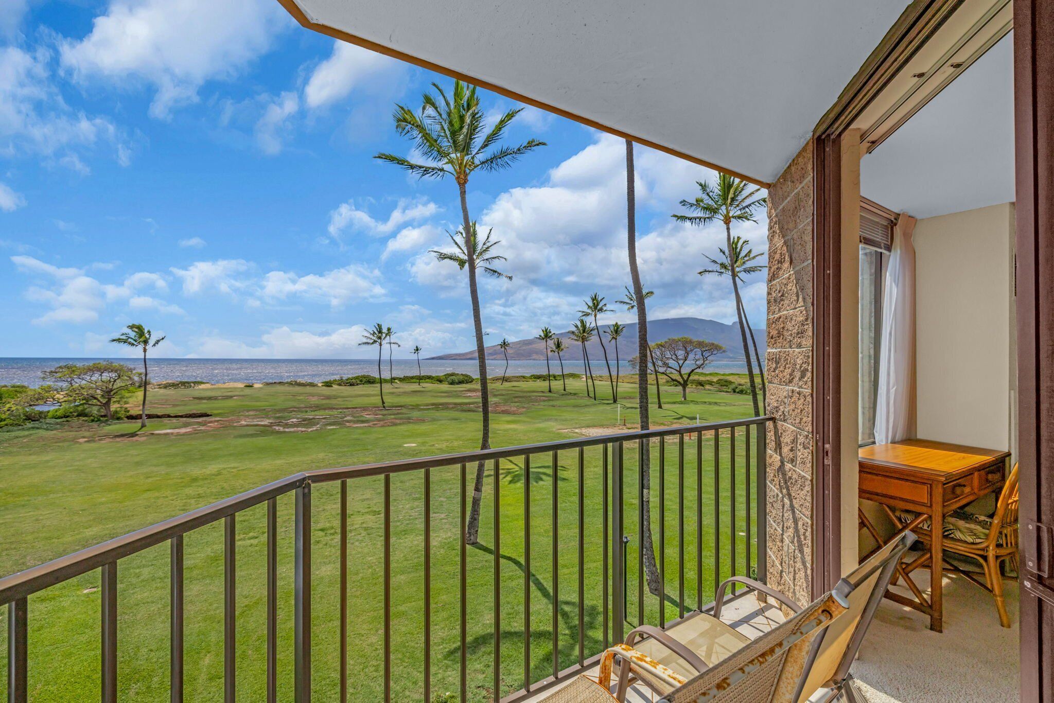 A balcony with a view of a grassy area and palm trees, a wooden table, and a chair.