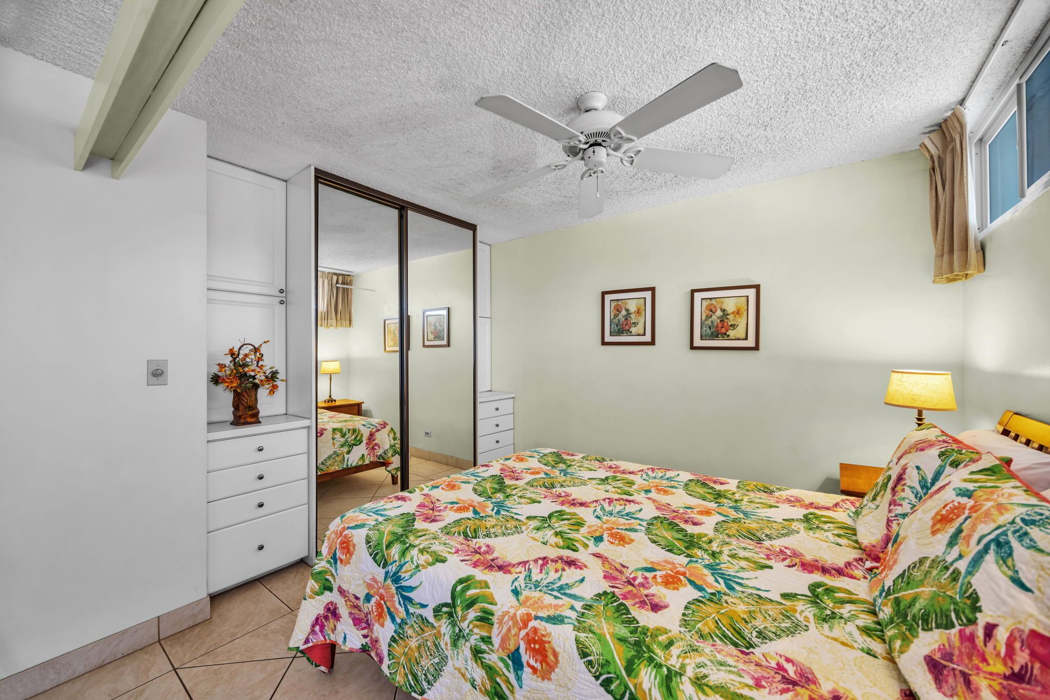 A bedroom with a bed, a ceiling fan, a dresser, a mirror, and two framed pictures on the wall.