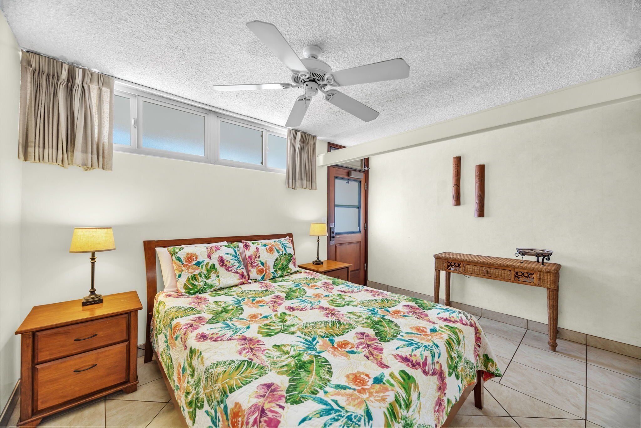 Une chambre à coucher comprenant un lit, une table de chevet, un bureau, un ventilateur de plafond et une fenêtre avec des rideaux.