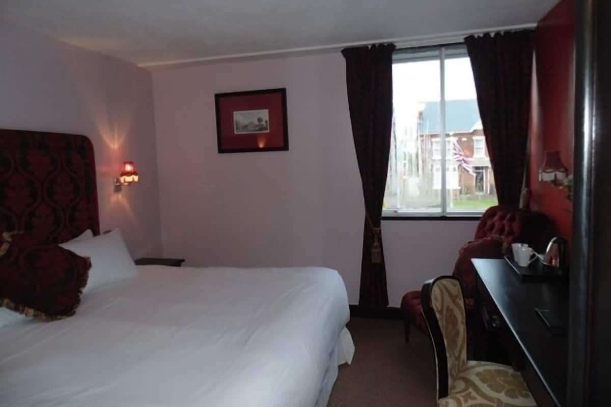 A hotel room with a large bed, a desk, a chair, a picture on the wall, and a window with curtains.