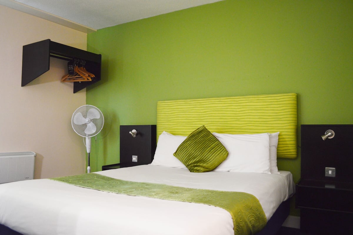 A neatly made bed with a green and white bedspread, a single green pillow, and a white fan beside the bed.
