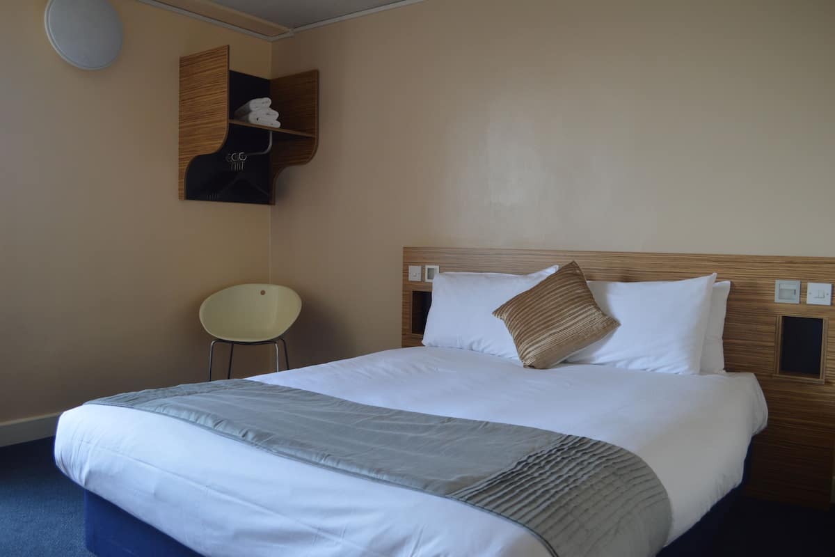 A neatly made bed with white linens and a grey throw blanket, a wooden headboard, a small white chair, and a wall-mounted shelf with towels.