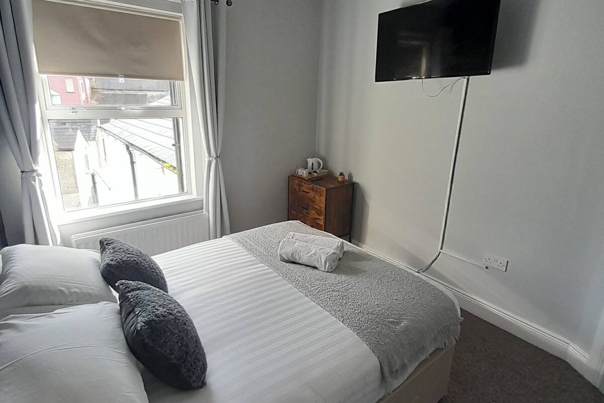 A bedroom with a bed, a TV mounted on the wall, a window with curtains, and a small wooden nightstand.