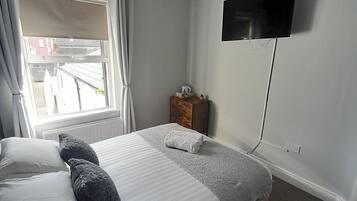 A bedroom with a bed, a TV mounted on the wall, a window with curtains, and a small wooden nightstand.