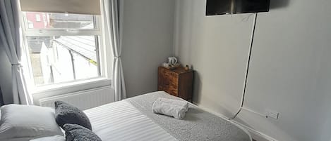 Un dormitorio con cama, televisor montado en la pared, ventana con cortinas y una mesita de noche de madera.