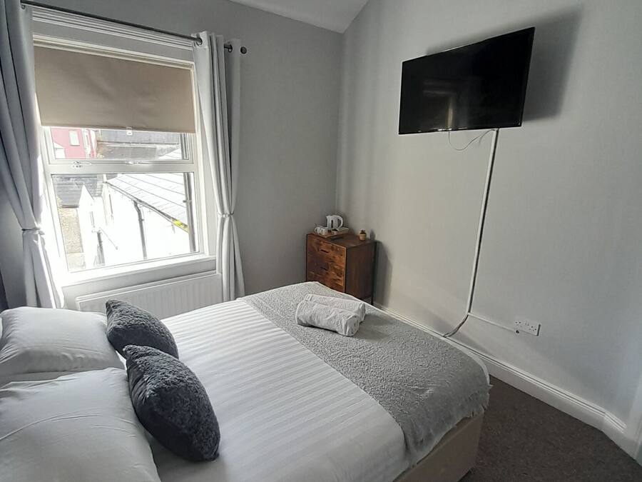 A bedroom with a bed, a TV mounted on the wall, a window with curtains, and a small wooden nightstand.
