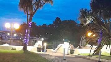 Un espacio exterior iluminado de noche, con palmeras, un camino y farolas.