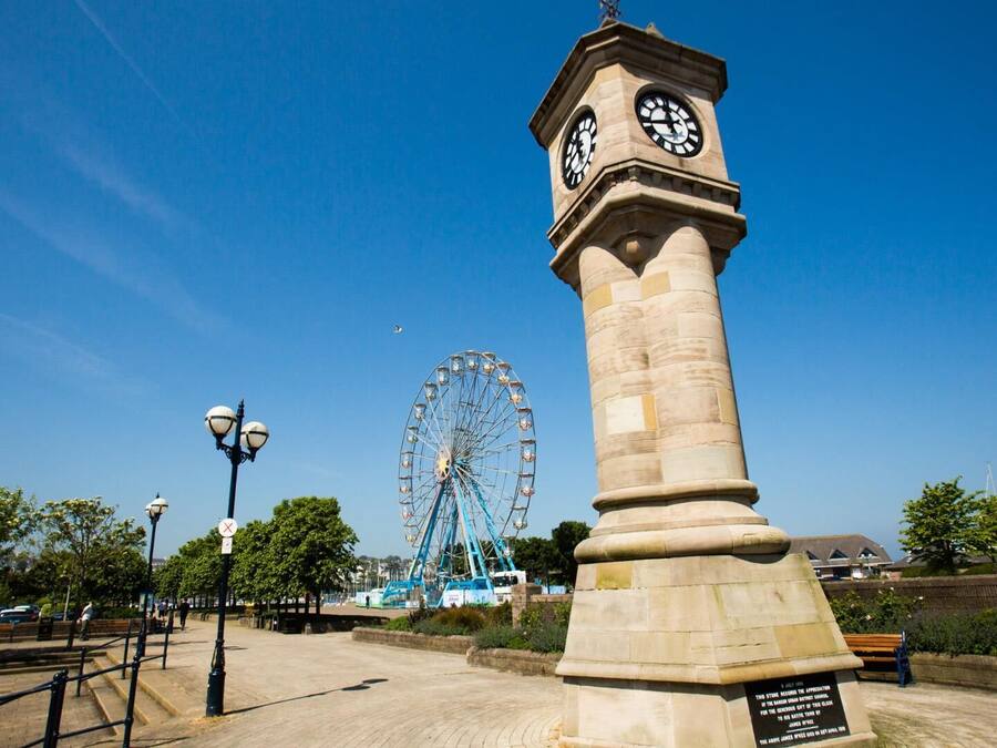 A clock tower with a plaque, a Ferris wheel, and a park with benches and lamp posts.