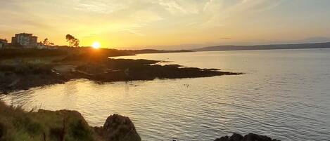 Un paisaje costero con un atardecer sobre el agua, costa rocosa y edificios a lo lejos.