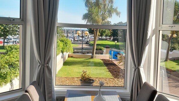 A dining area with a table set for two, large windows with curtains, a view of a landscaped area with a palm tree, and a distant view of a cityscape. - Astala Guesthouse (Bangor)