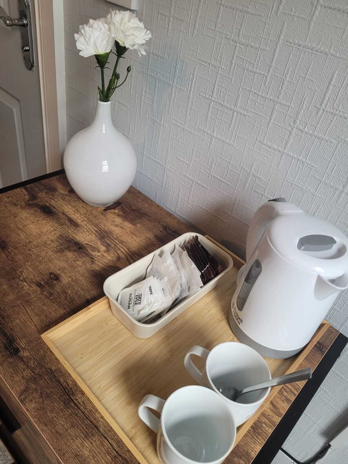 A wooden table with a white vase containing white flowers, a white teapot, a tray with sugar packets, and two white cups.