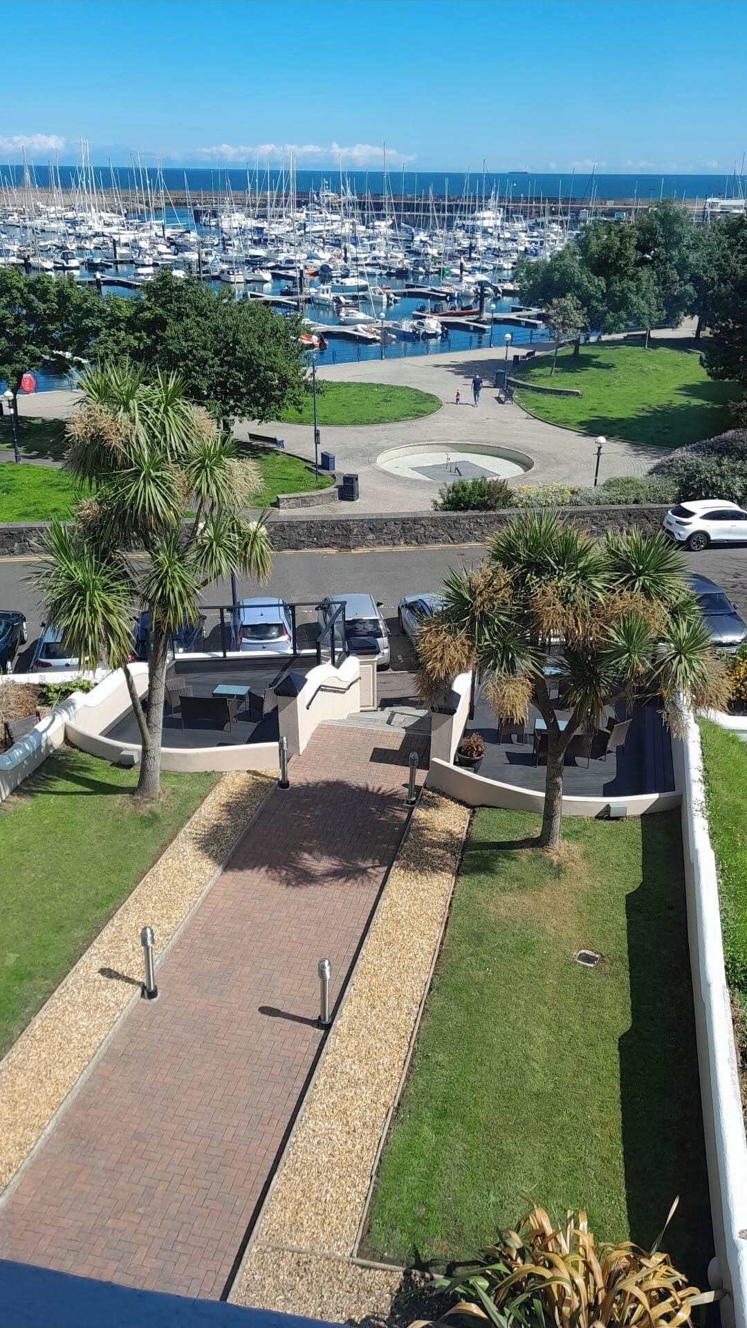 Un puerto deportivo con numerosas embarcaciones amarradas, un aparcamiento con coches y una zona ajardinada con palmeras.