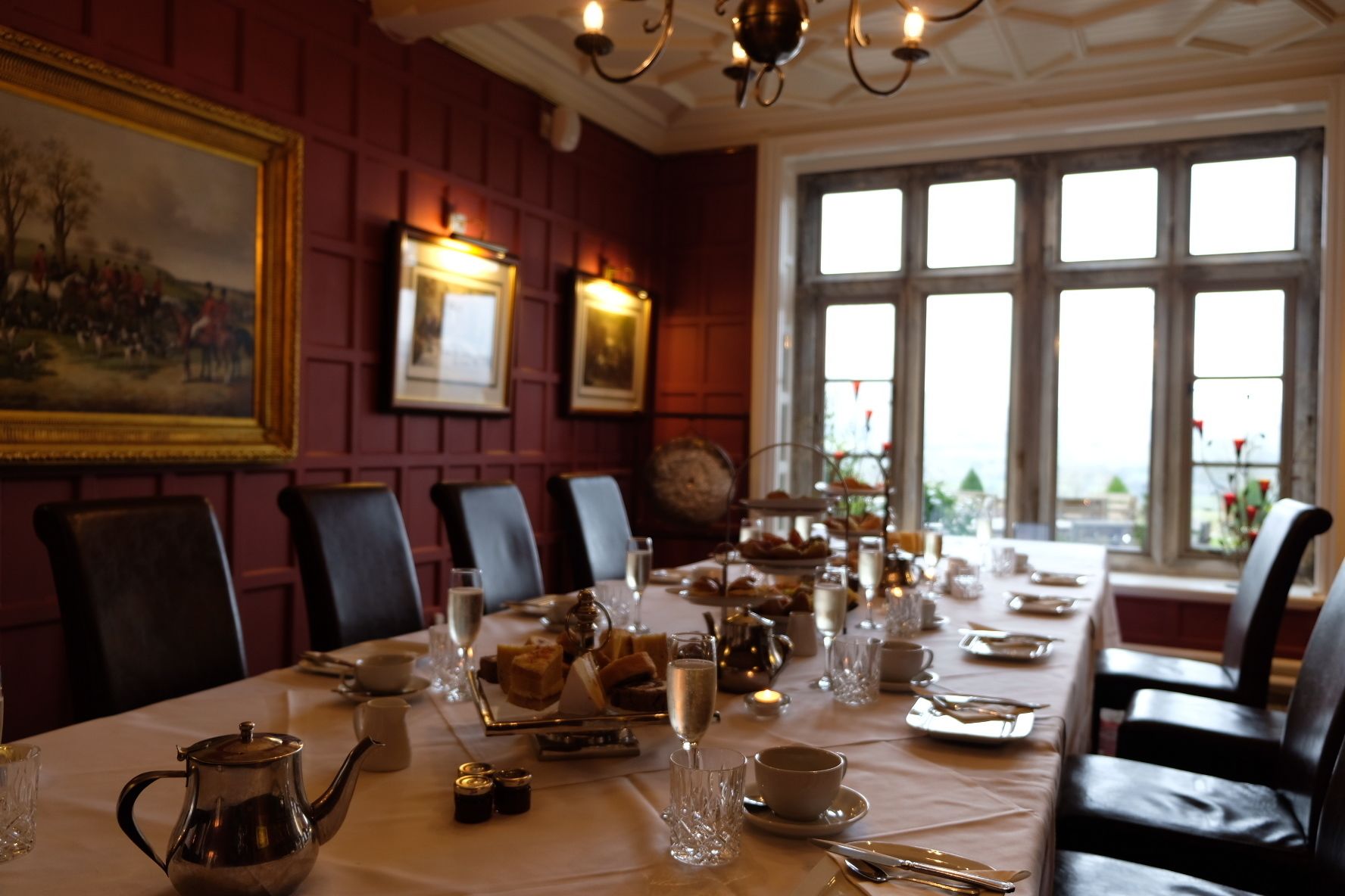 A dining room with a long table set for a meal, framed artwork on the walls, and a view through large windows.