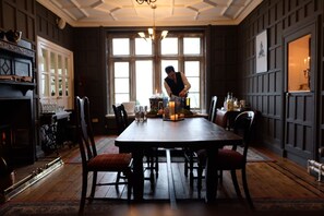 Ein Esszimmer mit großem Holztisch, Stühlen und einer Chandelier. Links befindet sich ein Kamin und ein Fenster mit vielen Glasscheiben.