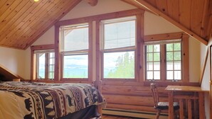 A cozy bedroom with a wooden ceiling, a large bed with patterned bedding, a wooden chair, and large windows offering a view of the outdoors. - Rustic 1 bedroom, 1 bath, 2 minutes from the West Glacier Park (West Glacier)