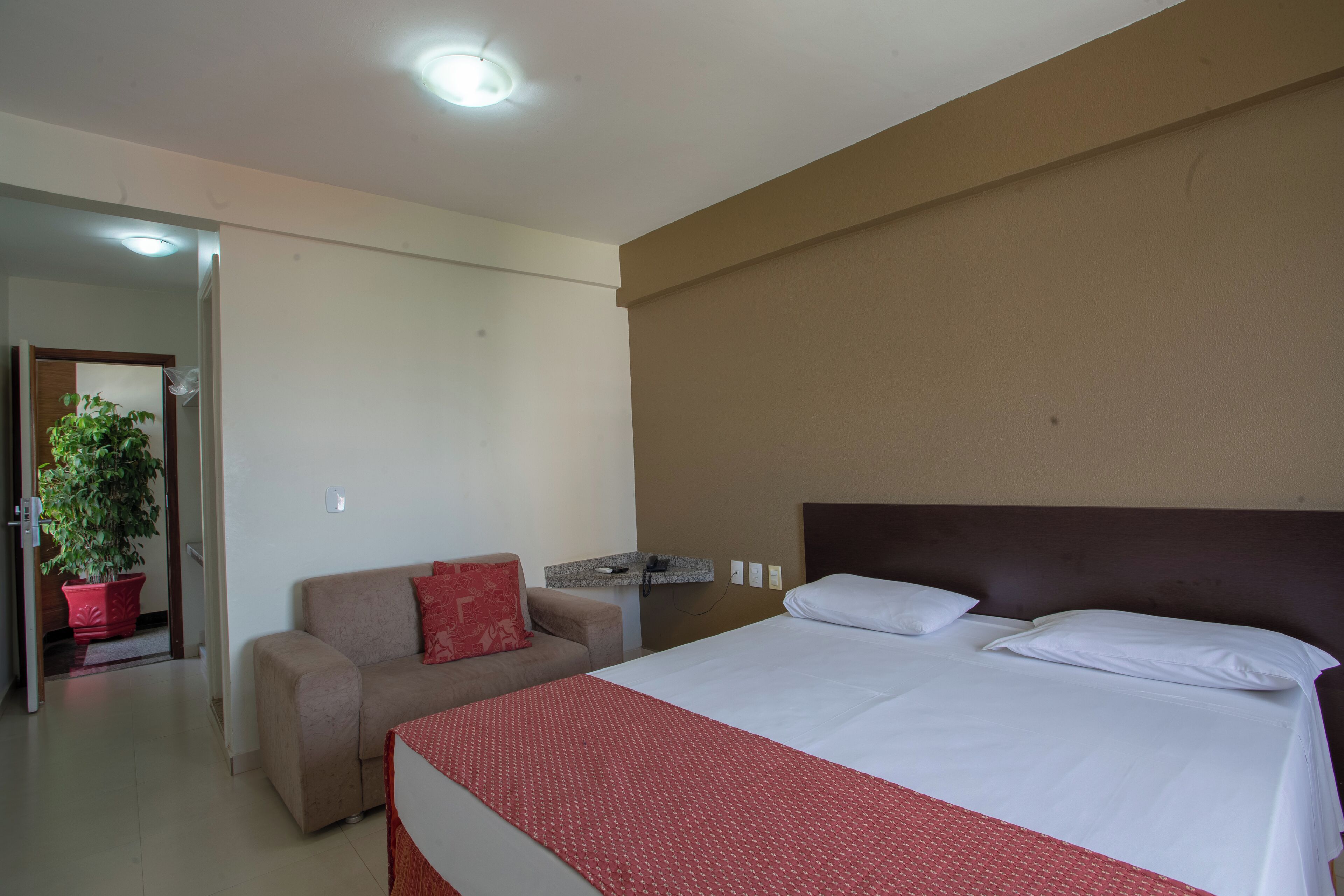 Quarto de hotel com cama, sofá, uma mesinha e um vaso de planta.