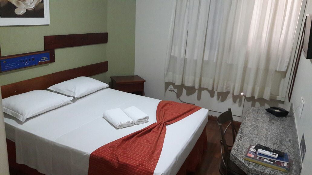 Habitación de hotel con cama, escritorio y silla, una mesita con libros y una cortina.