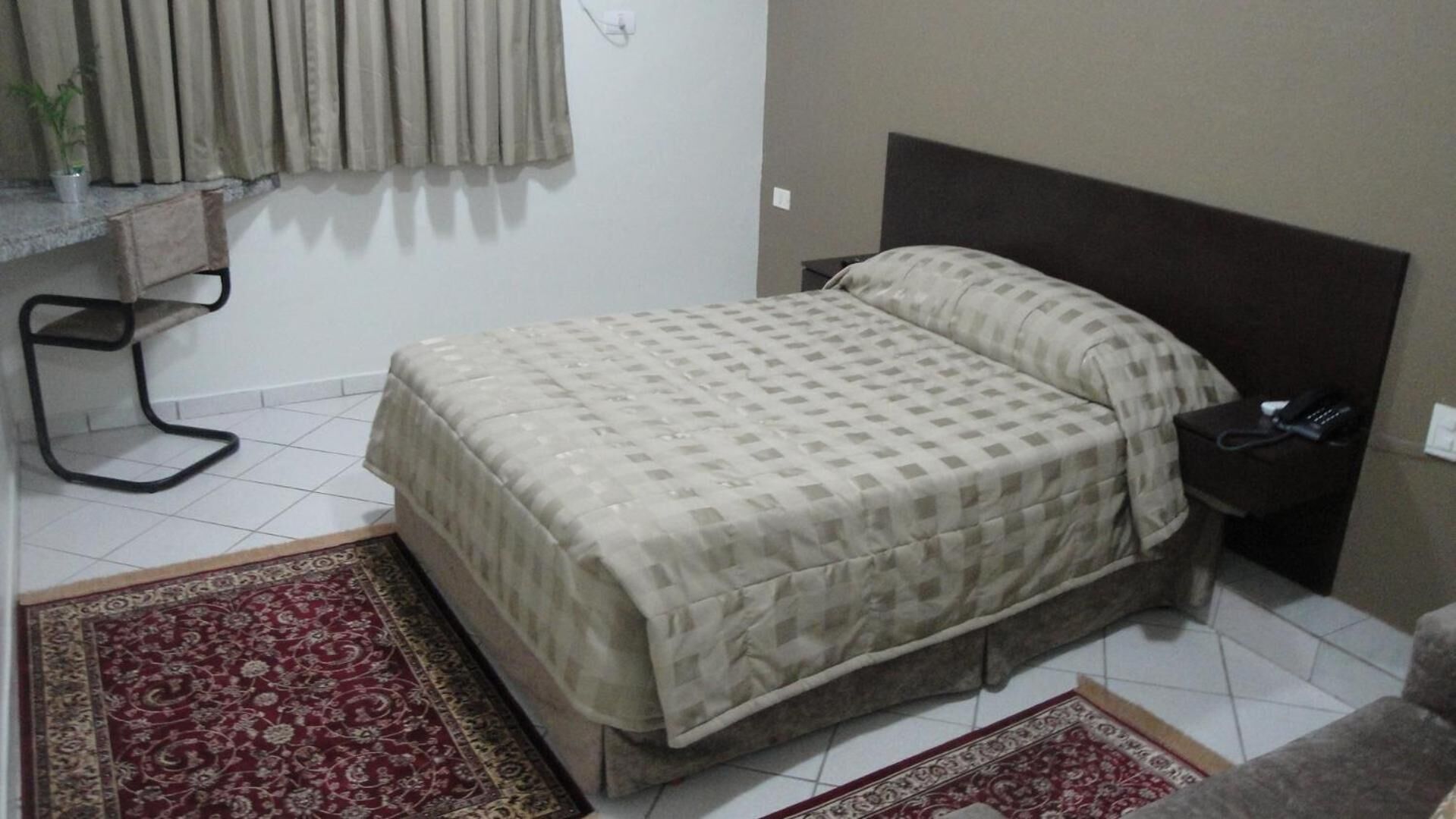 Un dormitorio con cama, mesita de noche, teléfono, silla y ventana con cortinas.