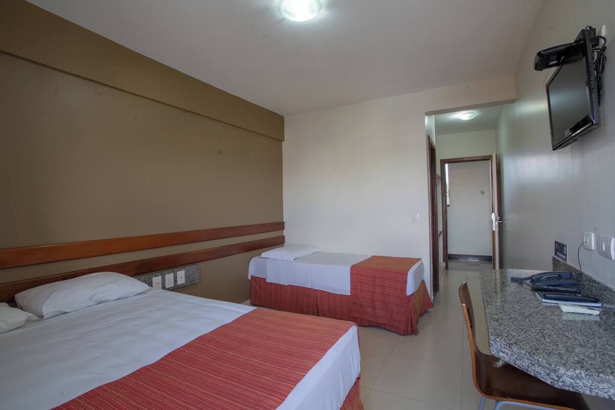 A hotel room with two beds, a desk, a chair, a TV mounted on the wall, and a door leading to another room.