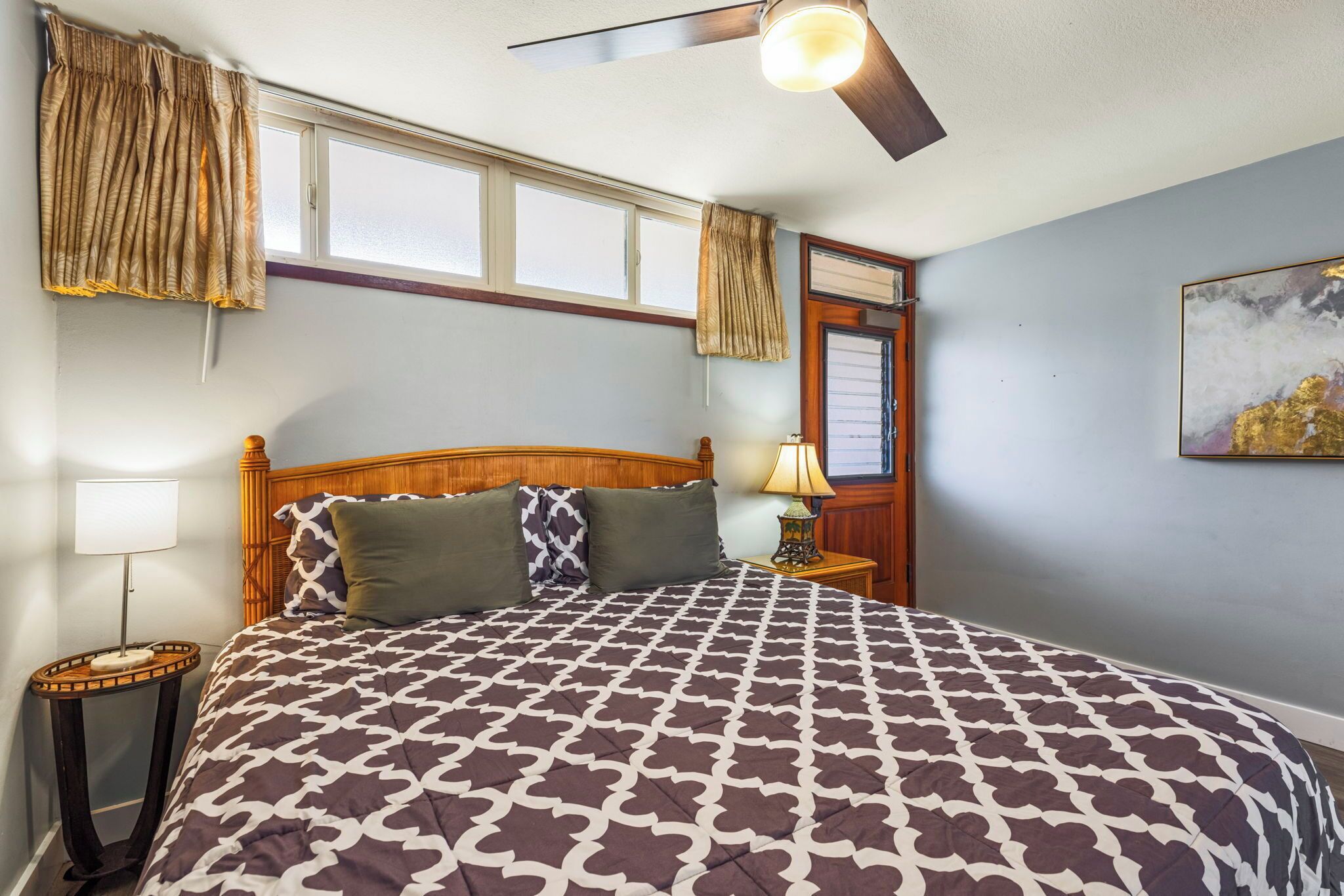 A bedroom with a large bed, a ceiling fan, a window with curtains, a nightstand with a lamp, and a painting on the wall.