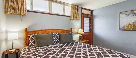 A bedroom with a large bed, a ceiling fan, a window with curtains, a nightstand with a lamp, and a painting on the wall.