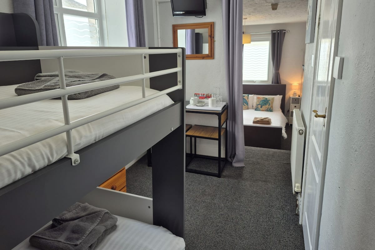 A bunk bed with a desk and a small table, a television mounted on the wall, and a window with curtains.