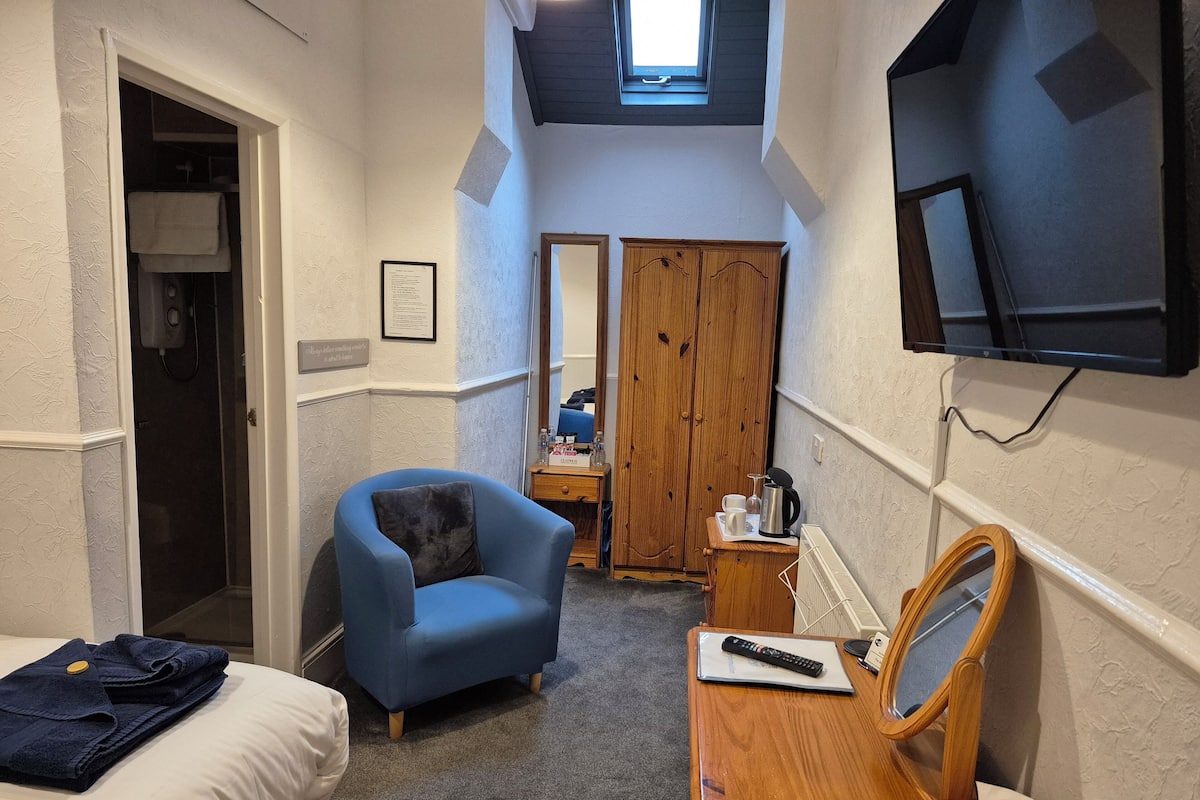 A hotel room with a bed, a blue armchair, a wooden dresser, a mirror, a television, and a door leading to a bathroom.