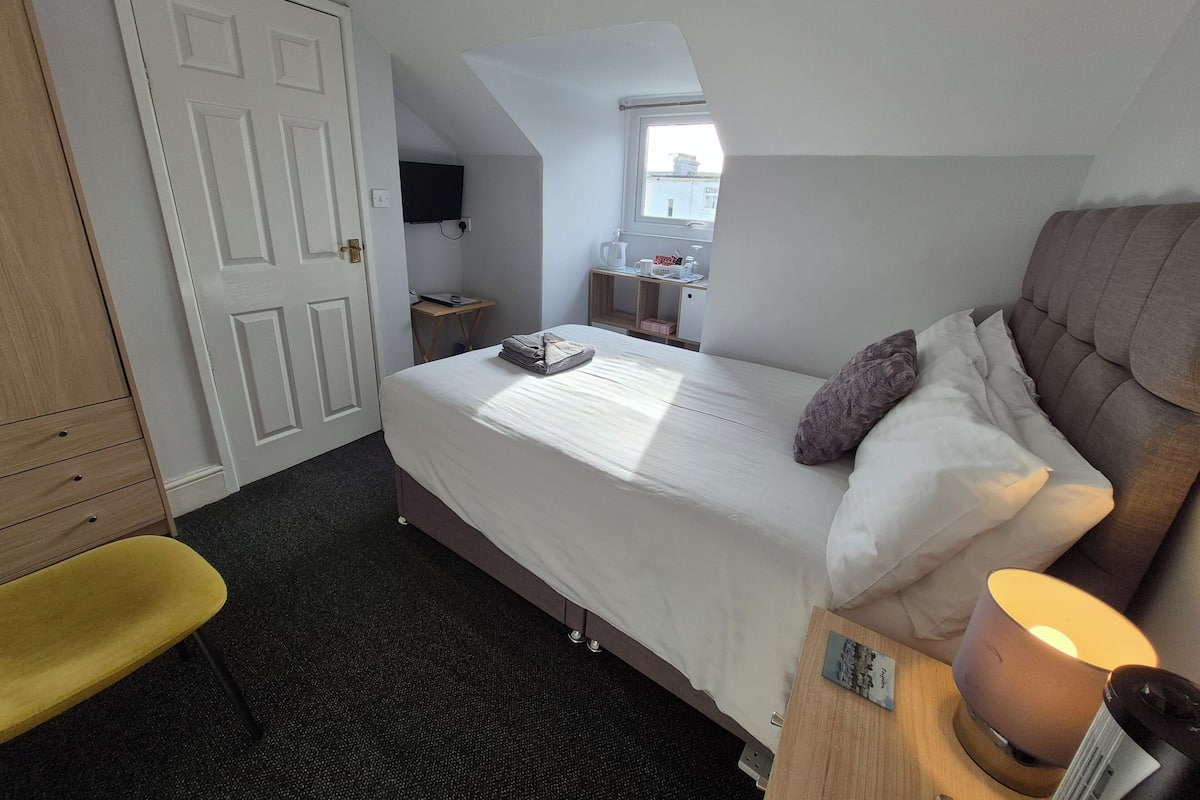 A bedroom with a bed, a desk with a lamp, a chair, a TV, and a window.