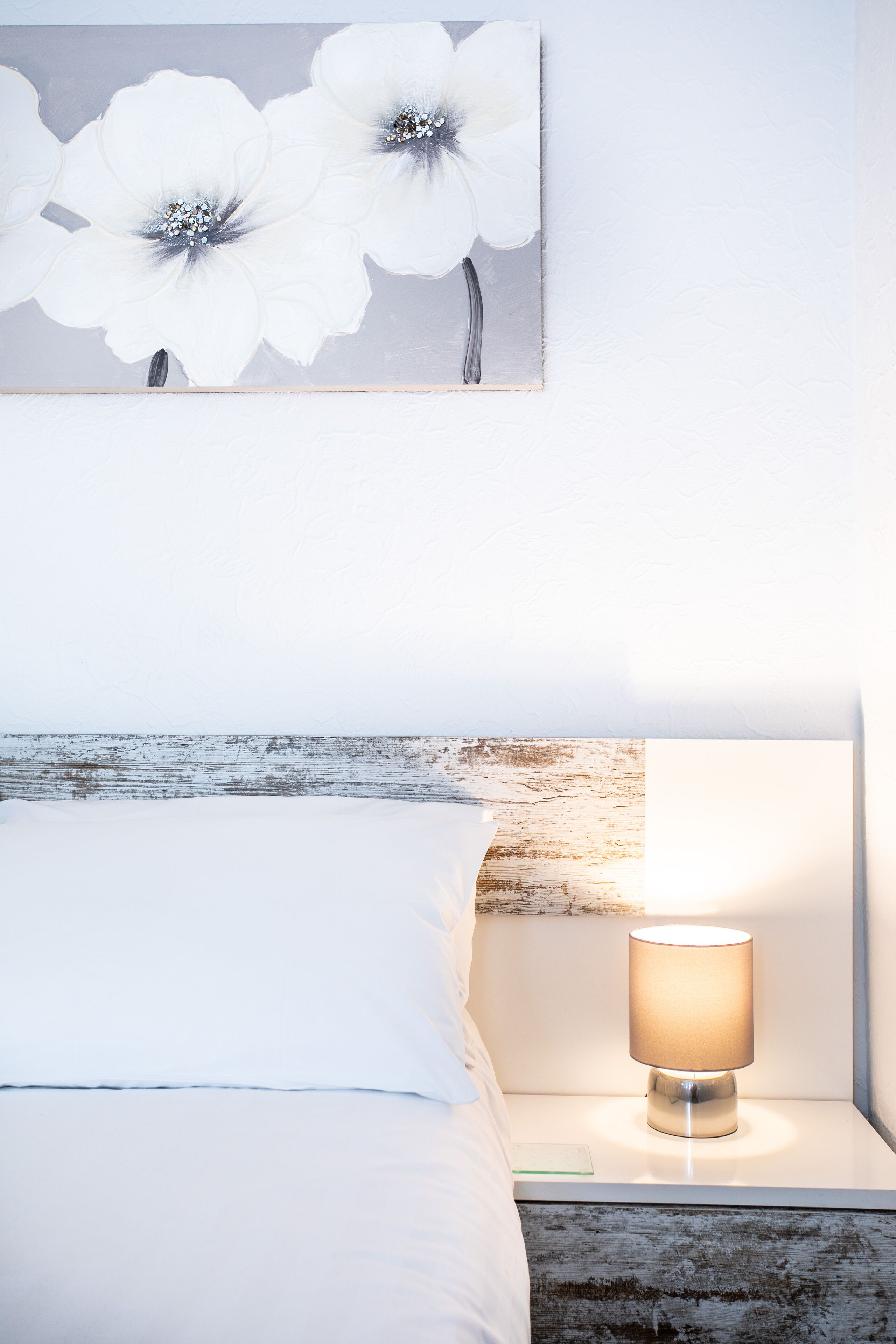 Una cama bien hecha con sábanas blancas, una lámpara de noche y un cuadro en la pared de flores blancas.
