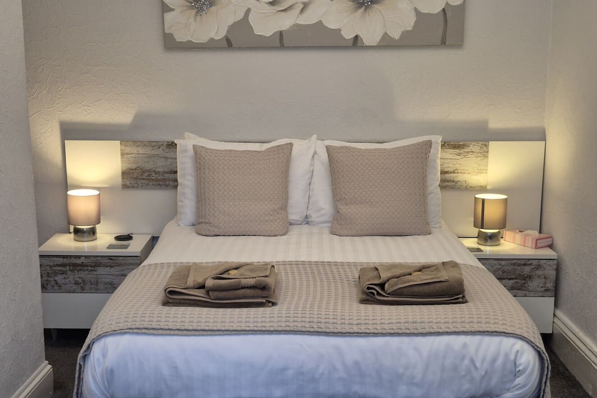 A neatly made bed with two pillows, a blanket, and two folded towels. There are two bedside tables with lamps and a small box on one.