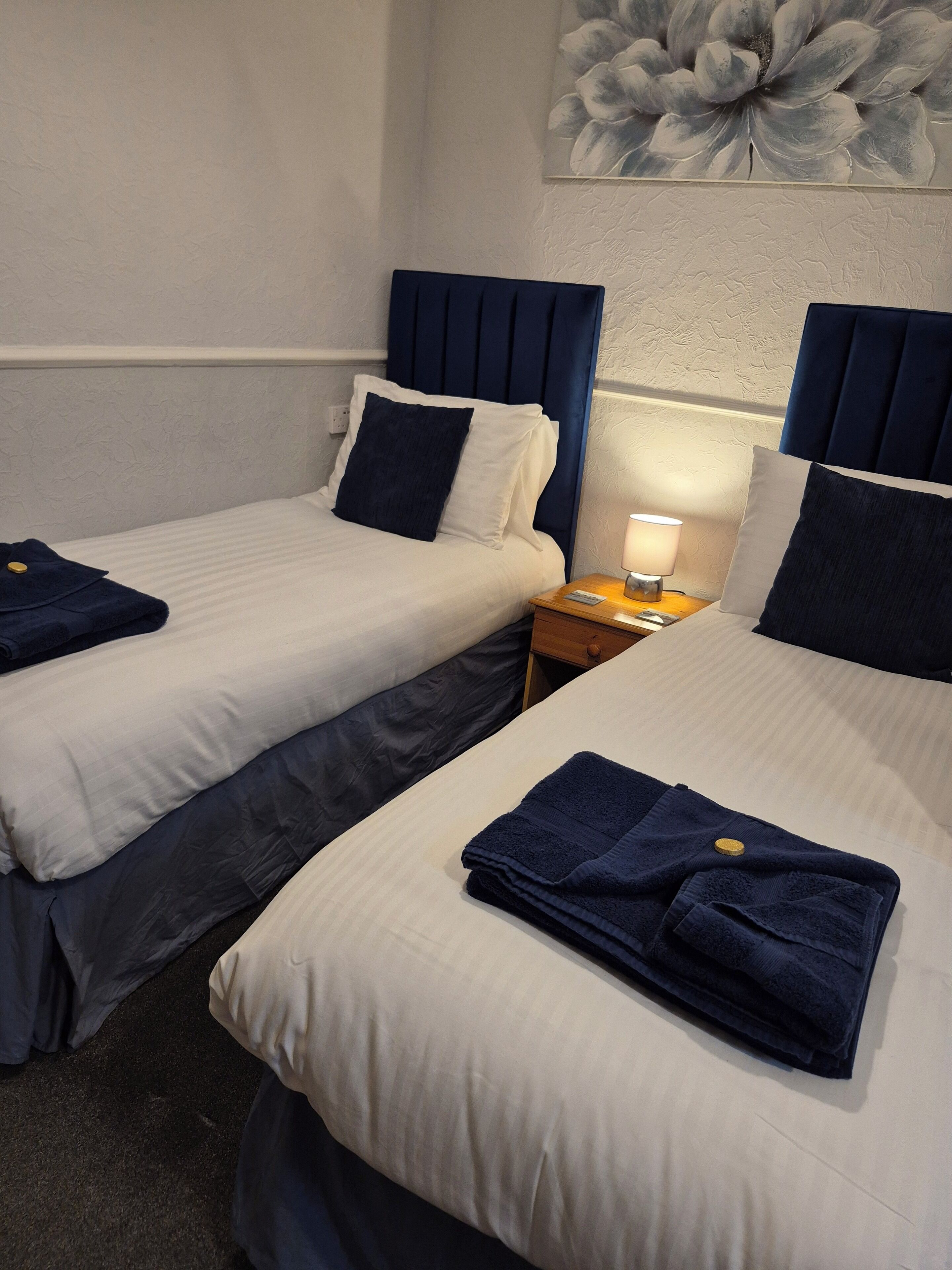 A hotel room with two beds, each with a blue headboard, white bedding, and a folded towel. There is a nightstand with a lamp and a small table between the beds.