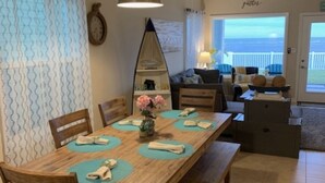 A dining area with a wooden table, chairs, and a bench. There is a mirror on the wall, a clock, and a view of the ocean through a door.