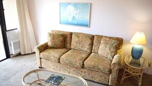 Una sala de estar con un sofá estampado de flores, una mesa de centro de vidrio con un libro, una lámpara y un cuadro en la pared.