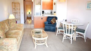 Una sala de estar con un sofá, una mesa de centro de vidrio, un sillón azul y una zona de comedor con sillas blancas y una mesa de vidrio.