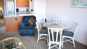 Un espacio habitable compacto con kitchenette, una zona de comedor con mesa de vidrio y sillas blancas, un sillón azul con cojín, y una lámpara colgante.